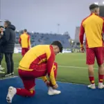 Calciatori in tuta giallorossa a bordo campo: uno si allaccia gli scarpini, un altro guarda il terreno di gioco