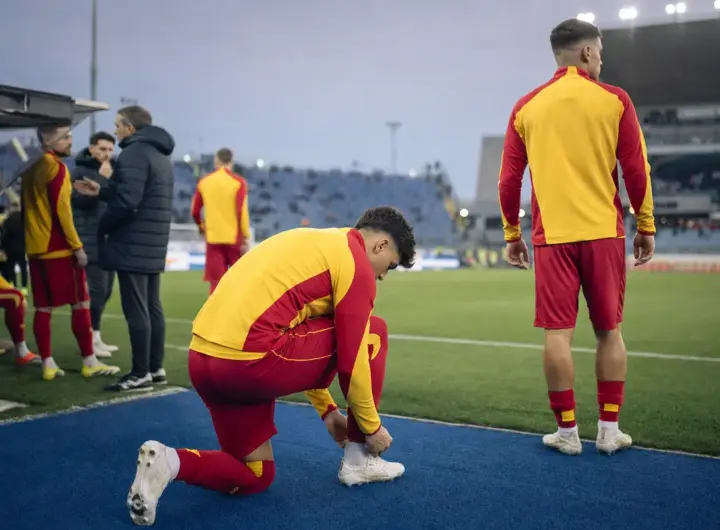 Calciatori in tuta giallorossa a bordo campo: uno si allaccia gli scarpini, un altro guarda il terreno di gioco