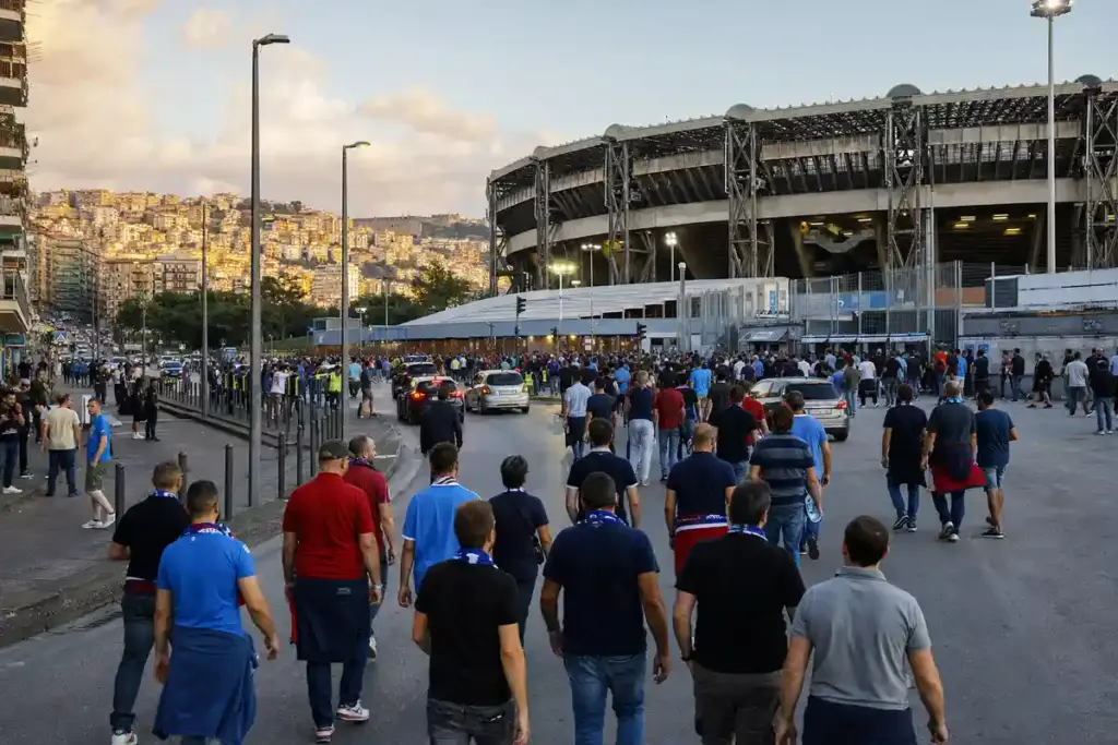 Tifosi in maglia azzurra e sciarpe camminano verso uno stadio al tramonto, con auto e transenne