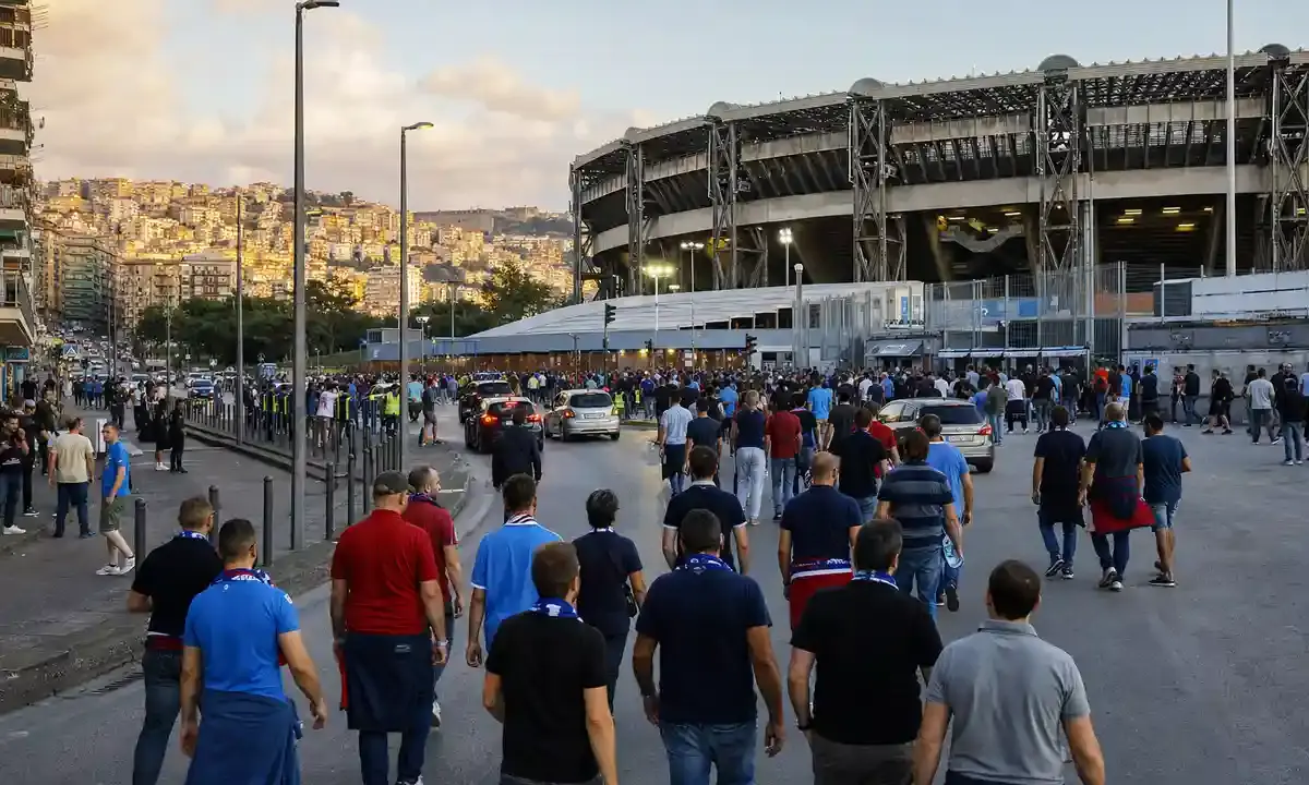 Tifosi in maglia azzurra e sciarpe camminano verso uno stadio al tramonto, con auto e transenne