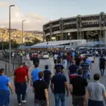Tifosi in maglia azzurra e sciarpe camminano verso uno stadio al tramonto, con auto e transenne