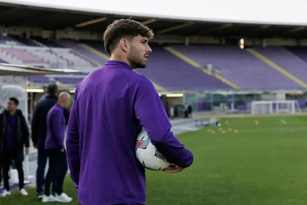 Calciatore in tuta viola con pallone sotto braccio a bordo campo, con tribune vuote sullo sfondo