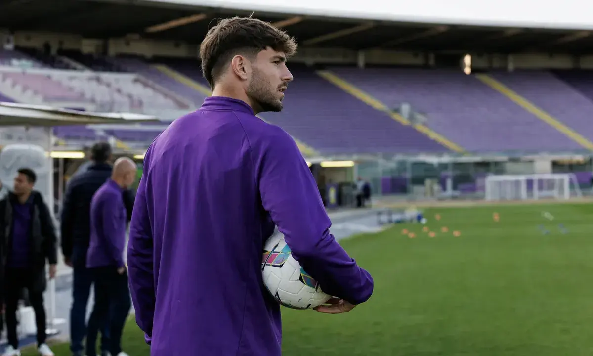 Calciatore in tuta viola con pallone sotto braccio a bordo campo, con tribune vuote sullo sfondo