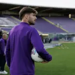 Calciatore in tuta viola con pallone sotto braccio a bordo campo, con tribune vuote sullo sfondo