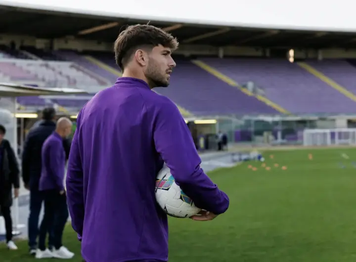 Calciatore in tuta viola con pallone sotto braccio a bordo campo, con tribune vuote sullo sfondo