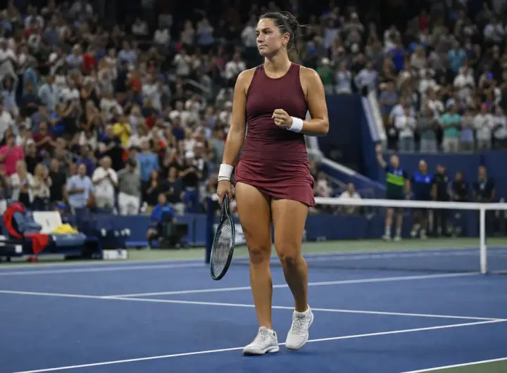 Tennista su campo in cemento blu, racchetta in mano e pugno chiuso, con il pubblico in piedi sugli spalti