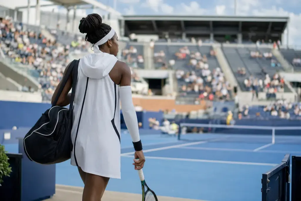 Tennista di spalle entra in uno stadio su campo blu con abito bianco con cappuccio, manicotto e borsa, racchetta in mano