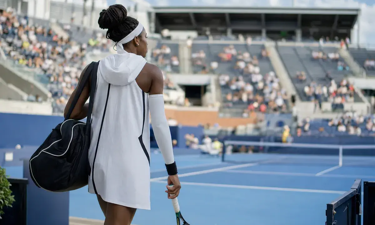 Tennista di spalle entra in uno stadio su campo blu con abito bianco con cappuccio, manicotto e borsa, racchetta in mano