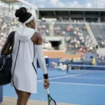 Tennista di spalle entra in uno stadio su campo blu con abito bianco con cappuccio, manicotto e borsa, racchetta in mano