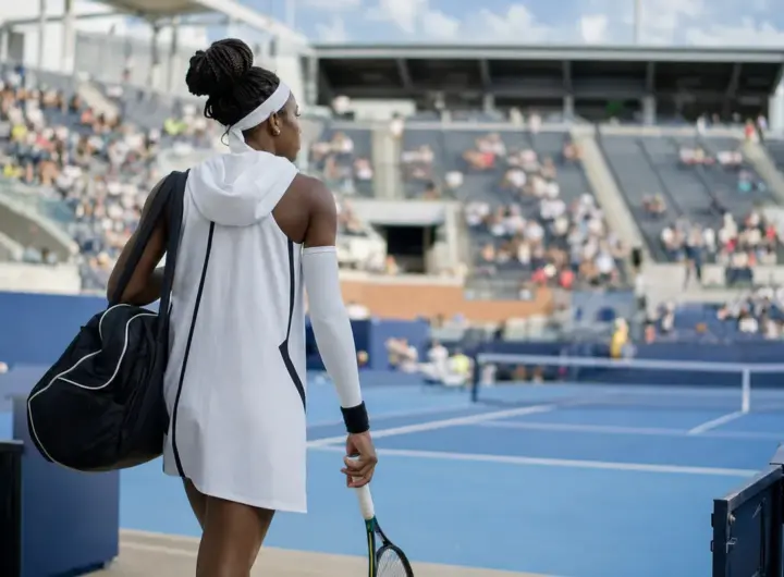 Tennista di spalle entra in uno stadio su campo blu con abito bianco con cappuccio, manicotto e borsa, racchetta in mano