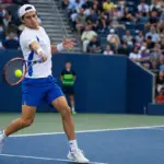 Tennista in maglia bianca e pantaloncini blu colpisce un diritto su un campo in cemento, con pubblico sugli spalti